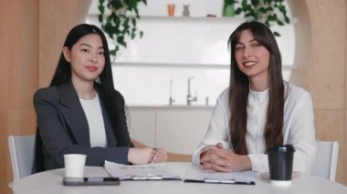 İki meslektaş, paket servis bardakları ve yazılı çizelgelerle küçük bir masada güven içinde oturuyorlar. Yüzü betimlenmiş ifadelerle öne dönük. Karşılaştıktan sonra gülümseyerek doğrudan kameraya bakmak..