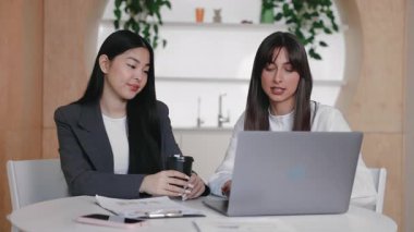 İki kadın profesyonel açık dizüstü bilgisayarla yan yana otururken projeyi tartışıyorlar. Biri kahve fincanını tutarken ve dikkatle dinlerken, diğeri ekrana işaret edip fikri açıklarken,
