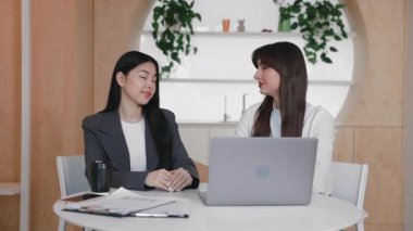 İki kendine güvenen iş kadını dizüstü bilgisayarları açık şekilde yan yana oturuyor ve masada kameraya bakıyor. Takım portresi için poz vermek veya sanal konferansa katılmak.