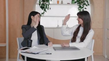 Yuvarlak masada gülerken ve öne eğilirken beşlik çakan iki mutlu profesyonel kadın. Parlak ve dostane ofis alanında kaynaşırken iş başarısını ya da başarılı fikri kutlamak.