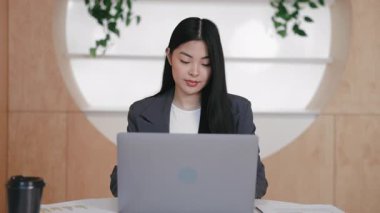 Konsantre Asyalı çalışan servis kahvesi ile masada otururken laptop ekranına bakıyor. İş e-postalarını okumak ya da performans sonuçlarını analiz etmek. Doğrudan kameraya bakıyorum..