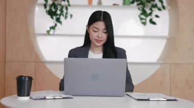 Profesyonel Asyalı kadın iki dosya ve kahve fincanı arasında otururken dizüstü bilgisayara odaklanmış. Parlak düzenli çalışma alanında yoğunlaşırken e- posta gözden geçiriliyor veya çevrimiçi konferansa katılılıyor.