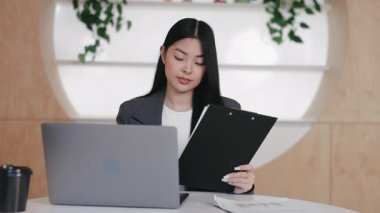 Analitik Asyalı ofis çalışanı açık dizüstü bilgisayarın yanında otururken panoda belge okuyor. İçeriği dikkatlice inceliyorum. Çevrimiçi tartışmadan önce içgörü hazırlanıyor veya rakamlar iki kez kontrol ediliyor.