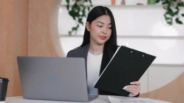Ciddi bir Asyalı kadın iş sırasında dizüstü bilgisayarın yanında otururken pano içeriğini kontrol ediyor. Rapor detayları doğrulanırken veya yazdırılan veriler dijital göreve devam etmeden önce gözden geçirilirken.