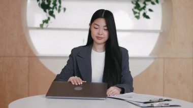 Düzenli ofiste uzak çalışma oturumuna hazırlanırken bilgisayarın kapağını kaldıran Asyalı kadın. Belgeler ve panonun yanında otururken sanal toplantıya hazırlanmak veya çevrimiçi görevlere başlamak.