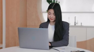 Güzel Asyalı yönetici dizüstü bilgisayarın arkasında oturuyor ve hafifçe gülümsüyor. Kamera için poz vermek veya profesyonel duruşu korurken video konferansına katılmak.