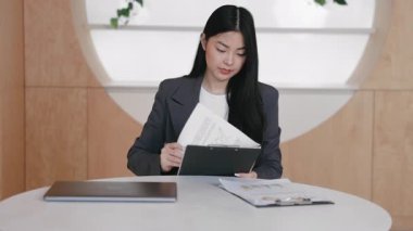 Masada otururken dosyasına iliştirilmiş belgelere göz atan kararlı Asyalı ofis çalışanı. Raporları analiz ederken veya toplantı notları hazırlarken çizelge ve iş verilerinin dikkatle incelenmesi.