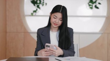 Sırıtkan Asyalı kadın bilgisayarlı ve yazılı çizelgelerle masada otururken akıllı telefona göz atıyor. Sosyal medyada neşeli mesajlar okumak ya da kısa aralarda güncellemeleri kontrol etmek. Uzak iletişim.