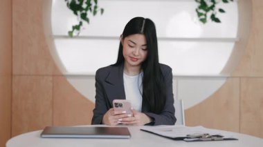 Mesajı okurken ya da yazarken iki elinle odaklanmış Asyalı profesyonel telefonu tut. Haritalar ve belgelerle çevrili modern çalışma alanında oturuyorlar. Siyah ceket giyiyor..