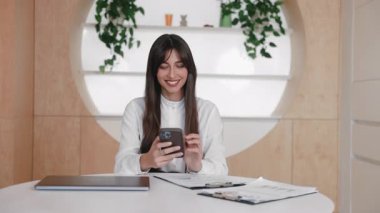 Memnuniyetle bayan ofis çalışanı telefona bakıyor ve ekranı kontrol ederken hafifçe gülüyor. Komik mesaja tepki vermek ya da masada otururken eğlenceli videolar izlemek. Belgelerle çevrili.