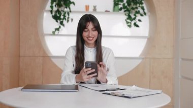 Şen şakrak esmer kadın iki eliyle akıllı telefonu tutarken düzenli çalışma alanında parlak bir şekilde gülümsüyor. Kapalı dizüstü bilgisayarın ve evrak işlerinin yanında oturuyor. Akıllı telefondan ileti okunuyor ya da uyarıya tepki veriliyor.