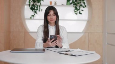 Kendine güveni tam bir kadın, temiz bir ofiste otururken akıllı telefon kullanıyor. Bilgisayarı ve panosu kapalı. Evrak işlerine kısa bir ara verirken akıllı telefondan mesaj yazmak ya da görüntüyü kaydırmak.