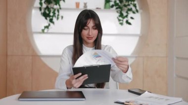 Zarif bir ofis çalışanı iş günü panoyu incelerken renkli rapor sayfalarını çeviriyor. Temiz örgütlü çalışma alanında kapalı dizüstü bilgisayarın yanında otururken istatistikler gözden geçiriliyor veya özet hazırlanıyor.