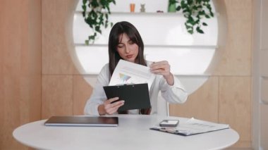 Güzel esmer kadın basılı sayfaları karıştırıyor ve düzenli bir masada otururken panodaki verileri kontrol ediyor. Rapor veya notları hazırlarken grafikler dikkatle gözden geçiriliyor.