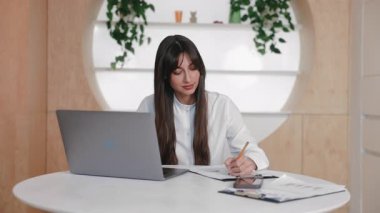 Ciddi bir esmer kadın, masa başında dizüstü bilgisayarla otururken evrak işlerine konsantre oluyor. Form dolduruluyor ya da rapora hazırlanırken aşağıya bakıyor ve temiz minimalist iç mekanda dikkatlice çalışıyor