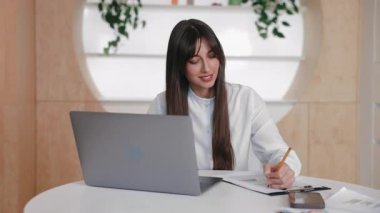 Barışçıl ofisteki laptop ekranına bakarken kağıda odaklanmış Kafkasyalı profesyonel yazı. Sanal sunum sırasında not almak veya uzak toplantıya katılırken verileri analiz etmek.