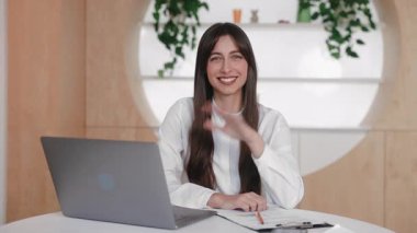 Çekici esmer kadın el sallarken modern çalışma alanında dizüstü bilgisayar ve belgelerle oturuyor. Video görüşmesi sırasında meslektaşınızı selamlamak ya da çevrimiçi konferans bittikten sonra veda etmek.