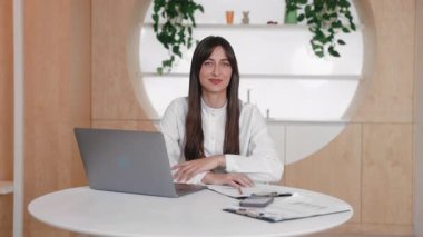 Pozitif beyaz kadın açık dizüstü bilgisayarın yanında oturuyor başparmağını kaldırıyor ve sırıtıyor. Onay ya da memnuniyet çevrimiçi toplantıyı tamamladıktan ya da uzak çalışma görevini başarıyla tamamladıktan sonra gösteriliyor