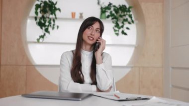 Elinde belgelerle masada otururken akıllı telefonu kulağına tutan sakin, uzun saçlı beyaz kadın. Gözlerini kapat ve konuşmaya odaklan. Önemli bir müşteri çağrısında dikkatle dinleniyor.