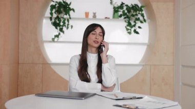 Modern masada oturan ve kollarını kavuştururken akıllı telefondan konuşan ciddi bir beyaz kadın. Proje tartışmak ya da iş görüşmesi yapmak. Sorunu çözmek veya ayrıntıları uzaktan görüşmek.