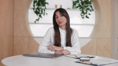 Odaklanmış zarif beyaz kadın yuvarlak masada oturuyor ve elleriyle hareket ederken kamerayla konuşuyor. Konuyu açıklamak ya da online sunum yapmak. Toplantı sırasında düşünceleri paylaşmak.