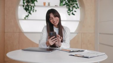 Beyaz masada otururken akıllı telefon ekranına parmaklarıyla bastıran beyaz bir kadın. Cevap yazılıyor ya da şakayla mesaj gönderiliyor. Arkadaşla sohbet etmek ya da internetteki fotoğrafa tepki vermek.