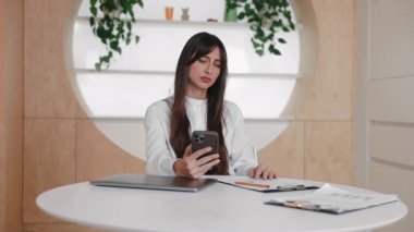 Beyaz masada oturan, yorgun bir ifadeyle akıllı telefonu tutan, hayal kırıklığına uğramış beyaz bir kadın. Sinir bozucu mesaj okumak ya da üzücü güncellemeleri kontrol etmek. Kötü haberleri işlerken sessizce tepki vermek.