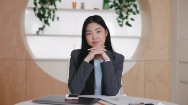 Kendine güvenen zarif Asyalı kadın sakince oturuyor elleri çenesinin altında katlanmış ve kameraya bakıyor. Önemli çevrimiçi toplantıdan önce bestelenmiş. Sanal tartışmada profesyonellik gösteriliyor.