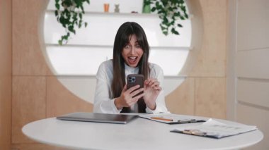 Beyaz masada otururken neşeli, beyaz bir kadın yumruğunu kaldırıyor ve elinde kocaman bir gülümsemeyle akıllı bir telefon tutuyor. Büyük başarıyı ya da online başarıyı kutluyoruz. Heyecan verici bir güncelleme aldıktan sonra gururlanmak.