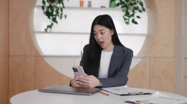 Ceketli sürpriz Asyalı kadın masada oturuyor ve geniş gözlü akıllı telefona bakıyor. Beklenmedik bir mesajı okuduktan sonra iki elle tutma cihazı. Şaşırtıcı güncellemeye duygusal tepki.