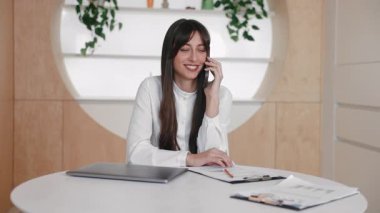 Elinde not defteri ve kağıtlarla masada otururken akıllı telefondan konuşan çekici, beyaz bir kadın. Keyifli bir görüşme ya da iyi haberler. Mola sırasında kalemi tutmak ve rahatlamak..