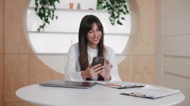 Şaşırmış esmer, beyaz kadın masada oturuyor ve iki eliyle akıllı telefon tutuyor. Mesaj okumak ya da heyecanla video izlemek. Sosyal medya beslemesine bakarken duygusal tepki.