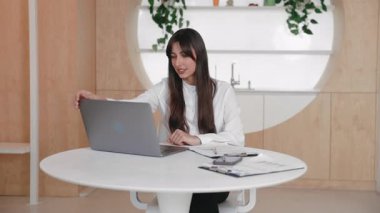 Pozitif uzun saçlı beyaz kadın masada oturuyor ve geniş bir gülümsemeyle doğrudan kameraya bakıyor. İlerlemeyi kutlarken sıkılmış yumrukları kaldırmak. Başarılı bir atışın ardından kendinden emin hissetmek.