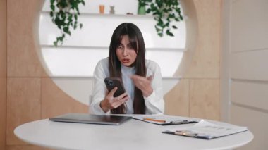 Sinirli beyaz kadın masada oturmuş akıllı telefonu tutuyor ve hayal kırıklığı içinde elini kaldırıyor. Video görüşmesi sırasında tartışmak ya da saldırı mesajına tepki vermek. Ekrana bakarken inançsızlığı ifade etmek.
