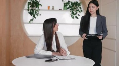 Spor ceketli, işine odaklanmış bir Asyalı kadın. Masaya yaslanmış, oturmakta olan meslektaşına evrakları gösteriyor. Strateji tartışmak ya da raporu birlikte gözden geçirmek. Ofis toplantısı sırasında etkin işbirliği.