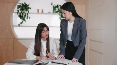 Endişeli Asyalı iş kadını panoya işaret ederken, sorunu vurgulayan Kafkasyalı meslektaşına açıklıyor. Oturan kadın hayal kırıklığı içinde başını tutuyor. Raporda acil durum tanımlanıyor.