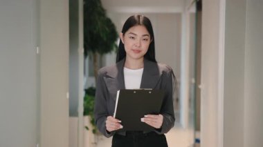 Spor ceketli kibar genç bir Asyalı kadın elinde bir pano ve modern koridorda kameraya bakıyor. Kendine güvenerek gülümsüyor. Takım toplantısında ya da mülakatta kendini tanıtmaya hazır..
