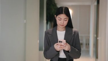 Resmi spor ceketli odaklanmış bir Asyalı kadın akıllı telefona bakıyor ve iki başparmağıyla da yazıyor. E- posta gönderiliyor ya da mesaja cevap veriliyor. Koridordaki dijital görevleri yönetirken konsantre olmak.