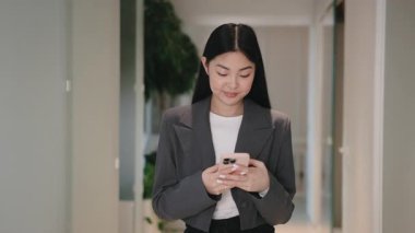 Gri ceketli şirin bir Asyalı kadın koridorda duruyor ve iki eliyle akıllı telefonu tutuyor. Nazikçe gülümsüyor. Yoğun ofis gününe hazırlanırken sabah güncellemeleri okunuyor ya da mesajlara cevap veriliyor.