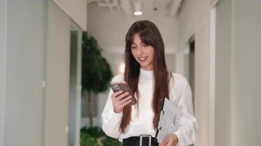Akıllı telefona bakıp koridorda yürürken gülümseyen uzun saçlı beyaz bir kadın. Komik mesajlar okumak ya da güncellemeleri kontrol etmek. Belgelerin tutulması ve iş günlerinde olumlu hisler