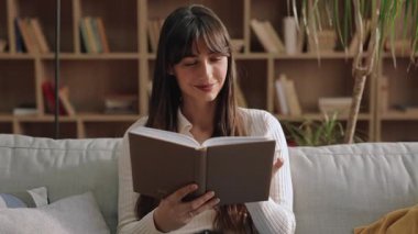 Gülümseyen beyaz kadın rahat bir kanepede oturuyor ve iki eliyle kitabı tutuyor. Dikkatlice okumak ya da ahşap kitaplığın yanındaki aydınlık odada huzurlu bir anın tadını çıkarmak..