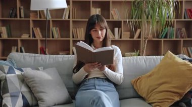 Kafkasyalı düşünceli bir kadın kucağında kitapla oturmuş başını kaldırırken çenesine dokunuyor. Hikaye üzerine düşünmek ya da kitaplarla dolu sessiz bir odada fikri analiz etmek ve yumuşak doğal ışık..