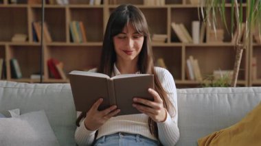 Güzel, beyaz bir kadın kanepede oturuyor ve kitap okurken elinde kitap tutuyor. Metne dalmak ya da arkada kitaplarla dolu ahşap raflarla sakin uzayda yeni bir şeyler öğrenmek..