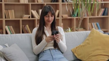 Çekici beyaz kadın iki eliyle akıllı telefon tutuyor ve koltukta otururken ışıl ışıl gülümsüyor. Kitaplarla dolu sıcak bir odada mutlu olmak ya da sohbetten zevk almak..