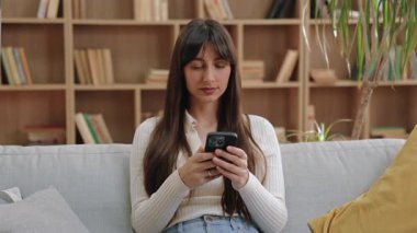 Kanepede hareketsiz oturan ve iki eliyle akıllı telefon tutan ciddi odaklı beyaz kadın. Kitap raflarıyla dolu sessiz bir odada ileriye bakıyor ya da dikkatle dinliyor..