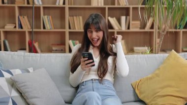 Akıllı telefonu tutarak, sevinçle yumruğunu kaldırarak ve şakacı bir şekilde dilini dışarı çıkararak kutlayan heyecanlı beyaz kadın. Işıkla dolu bir odada, arka planda kitap dolu raflarla kanepede oturmak..