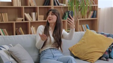 Gülümseyen beyaz bir kadın kanepeye oturmuş akıllı telefonu tutuyor ve el sallıyor. Yastıklar ve arkadaki ahşap kitaplık ile aydınlık, rahat bir odada arkadaşla görüntülü konuşma yapmak..