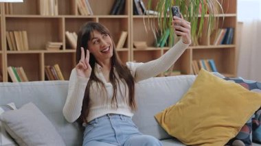 Çekici beyaz kadın kanepede oturmuş, akıllı telefona gülümserken parmaklarını barış işaretinde kaldırıyor. Kitap rafı ve yastıkları olan neşeli bir odada selfie çekmek veya video kaydetmek.