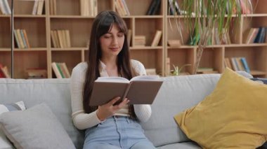 Kanepede oturup kitap okuyarak sayfayı çeviren güzel, beyaz bir kadın. Sessiz anın tadını çıkarmak ya da kitaplarla dolu yumuşak yastıklar ve raflarla huzurlu bir odada içeriği incelemek..