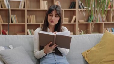 Gülümseyen beyaz kadın kütüphanede oturmuş elinde açık bir kitapla kameraya bakıyor. Huzurlu bir anın tadını çıkarmak ya da kitaplarla dolu ahşap raflarla çevrili olarak çalışmaya hazırlanmak..